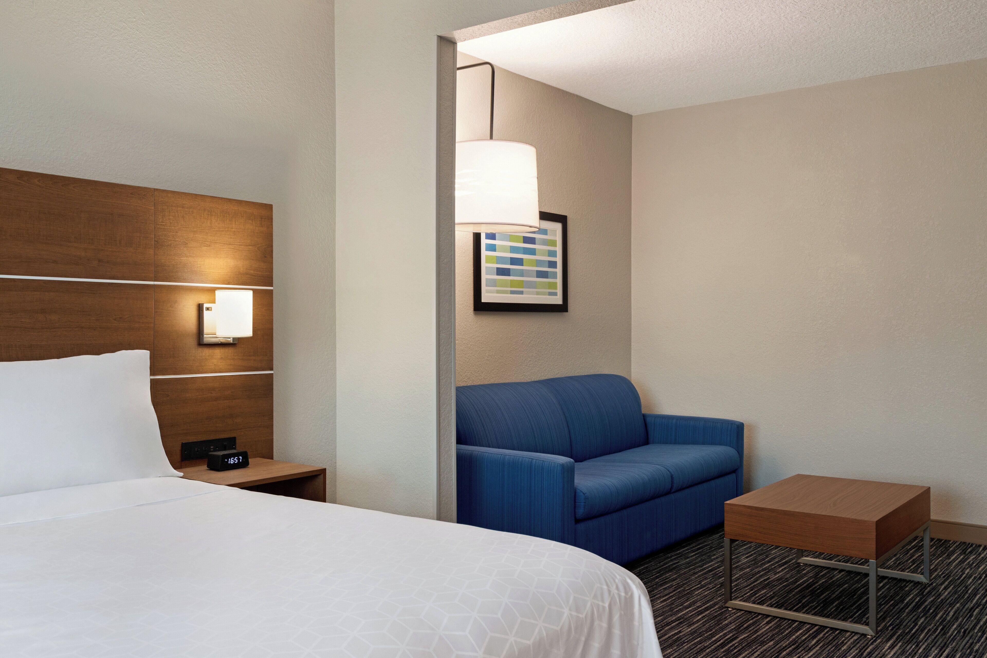 A hotel room with a bed, a nightstand, a blue sofa, a small table, and a wall-mounted lamp.