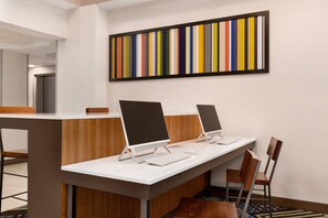 Uma área de recepção de hotel moderna, com três computadores de mesa, um balcão de madeira e uma pintura abstrata colorida na parede.