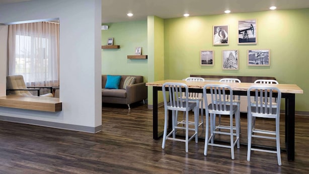 A modern dining area with a wooden table and metal chairs, a sofa with a blue pillow, and a wall with framed pictures. - WoodSpring Suites McKinney (McKinney)