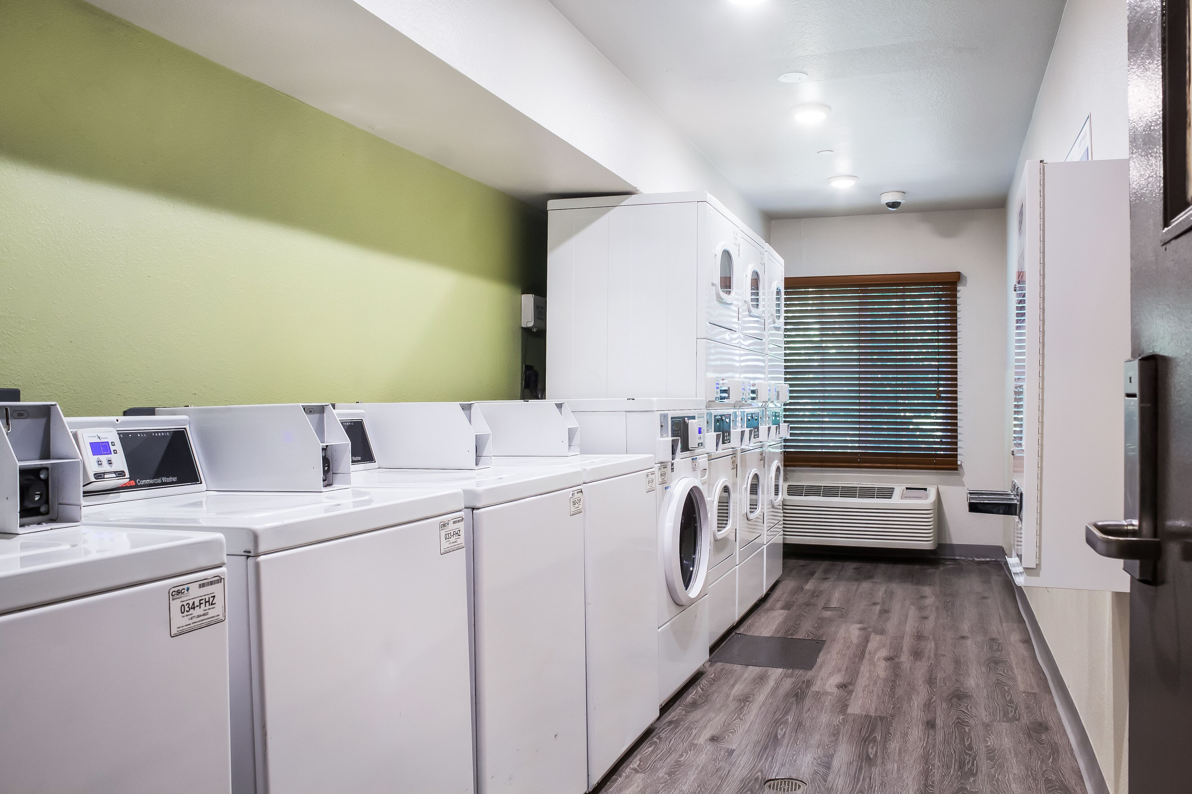 A laundry room with multiple washing machines and dryers, a control panel, and a window with blinds.