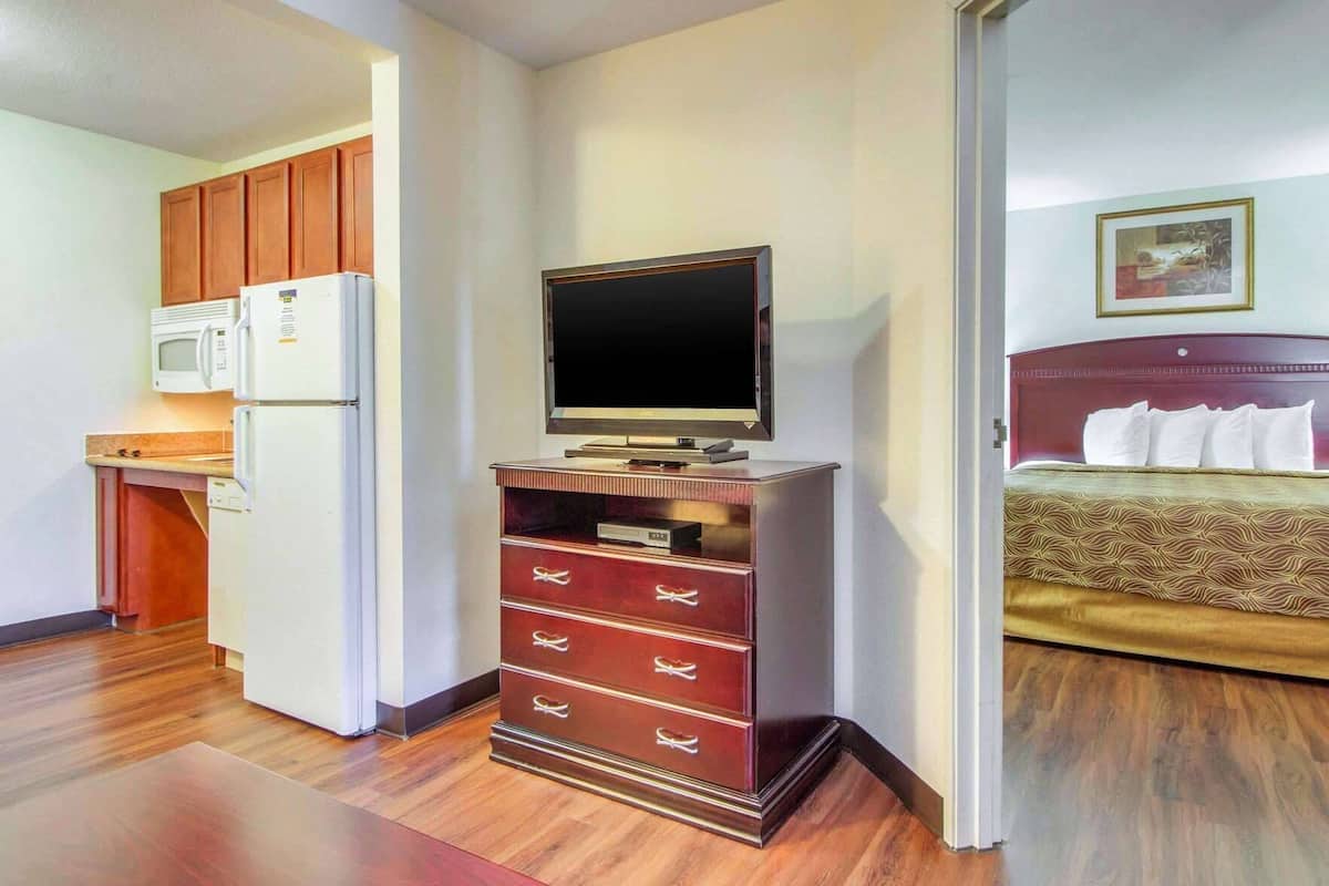 Een hotelkamer met een kitchenette, een televisie op een houten standaard, een bed met een geborduurd sprei en een ingelijst schilderij aan de muur.