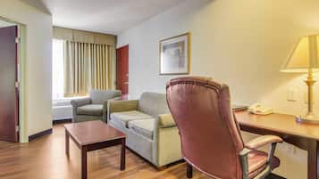 Habitación de hotel con piso de madera, un sofá, una silla, una mesita, una lámpara y un cuadro en la pared.