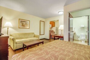 A hotel room with a bed, a sofa, a desk, a chair, a lamp, a picture on the wall, and a bathroom visible through a glass door. - MainStay Suites Texas Medical Center/Reliant Park (Houston)