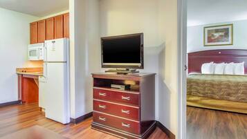 A hotel room with a kitchenette, a television on a stand, a bed with a headboard, and a painting on the wall.