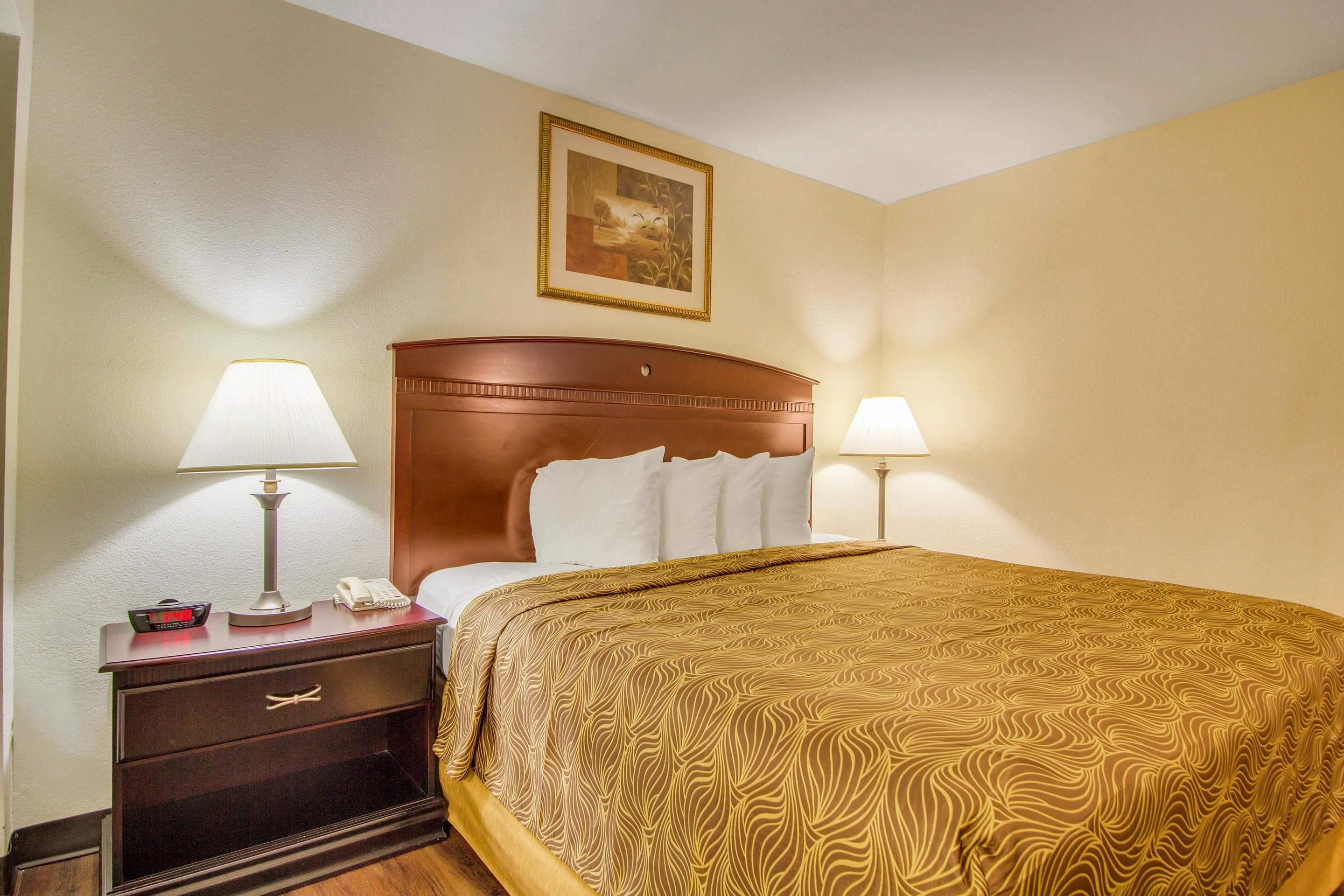 A hotel room with a bed, bedside lamps, a nightstand with a phone, a drawer, and a painting on the wall.