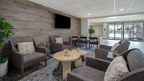 A modern lobby with grey upholstered seating, a wooden accent wall, a flat-screen TV, and a patterned carpet.