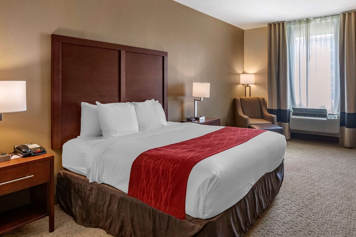 A hotel room with a large bed, a wooden headboard, a desk with a telephone, a chair, and a window with curtains.