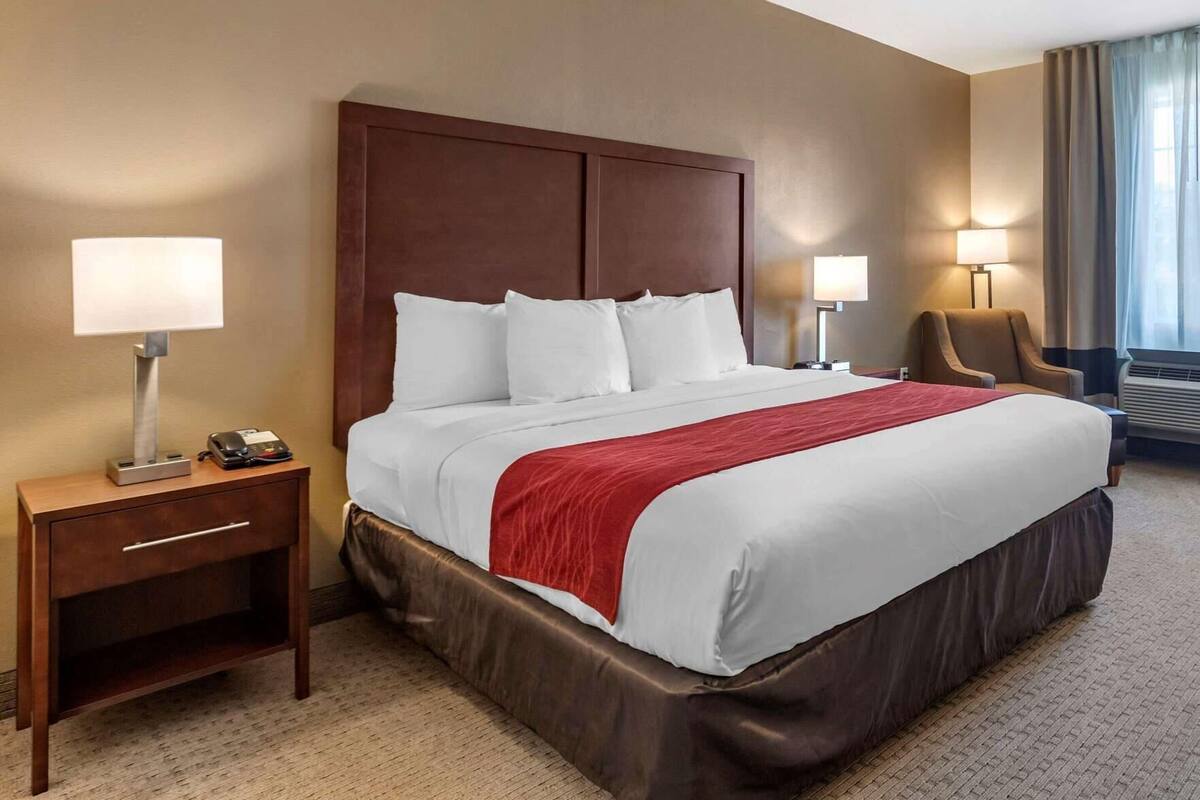 A hotel room with a large bed, a nightstand with a lamp, a chair, and a desk with a phone.