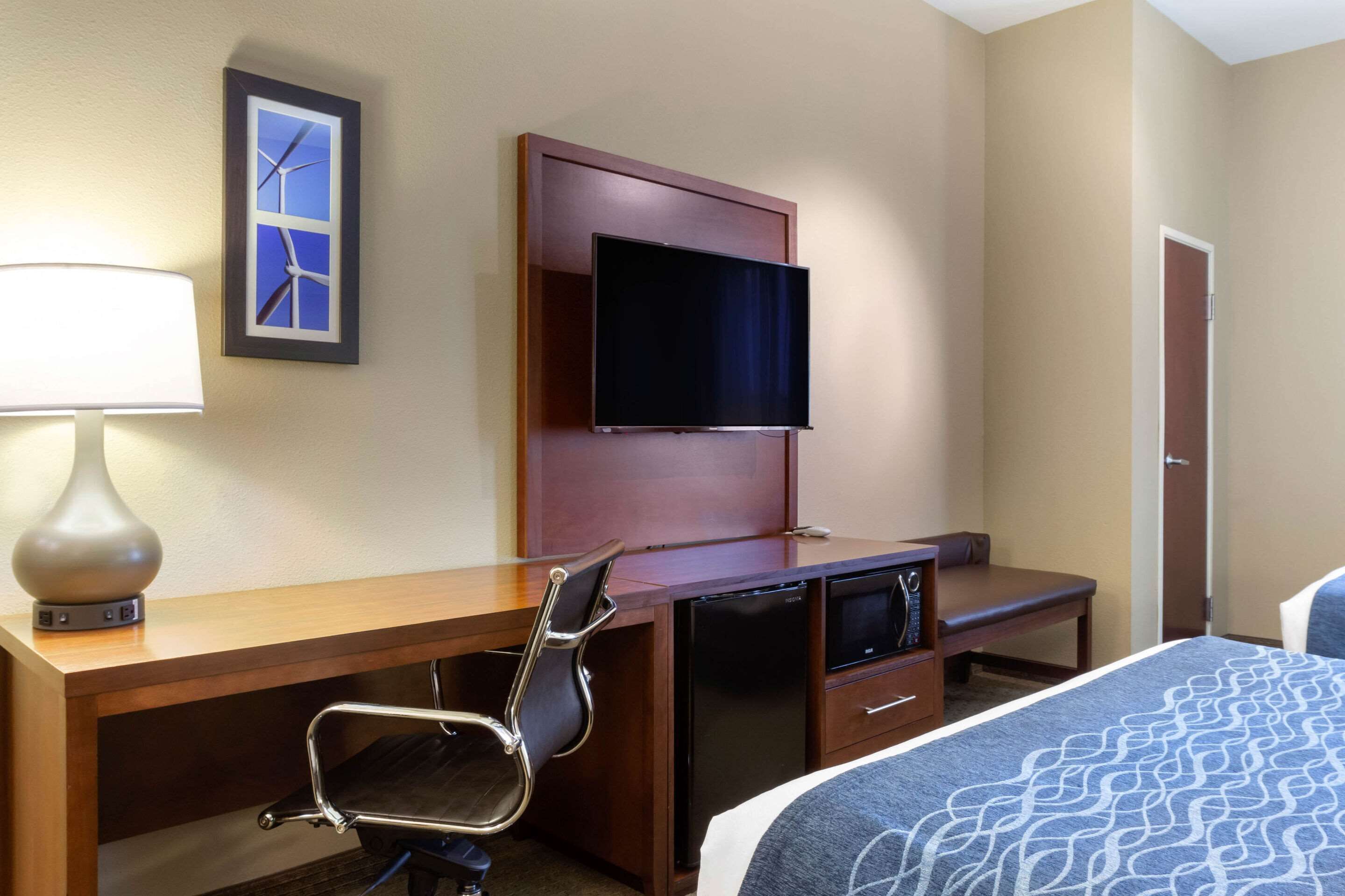 A hotel room with a wooden desk, a television mounted on the wall, a chair, a bed with a patterned bedspread, and a framed picture of wind turbines.