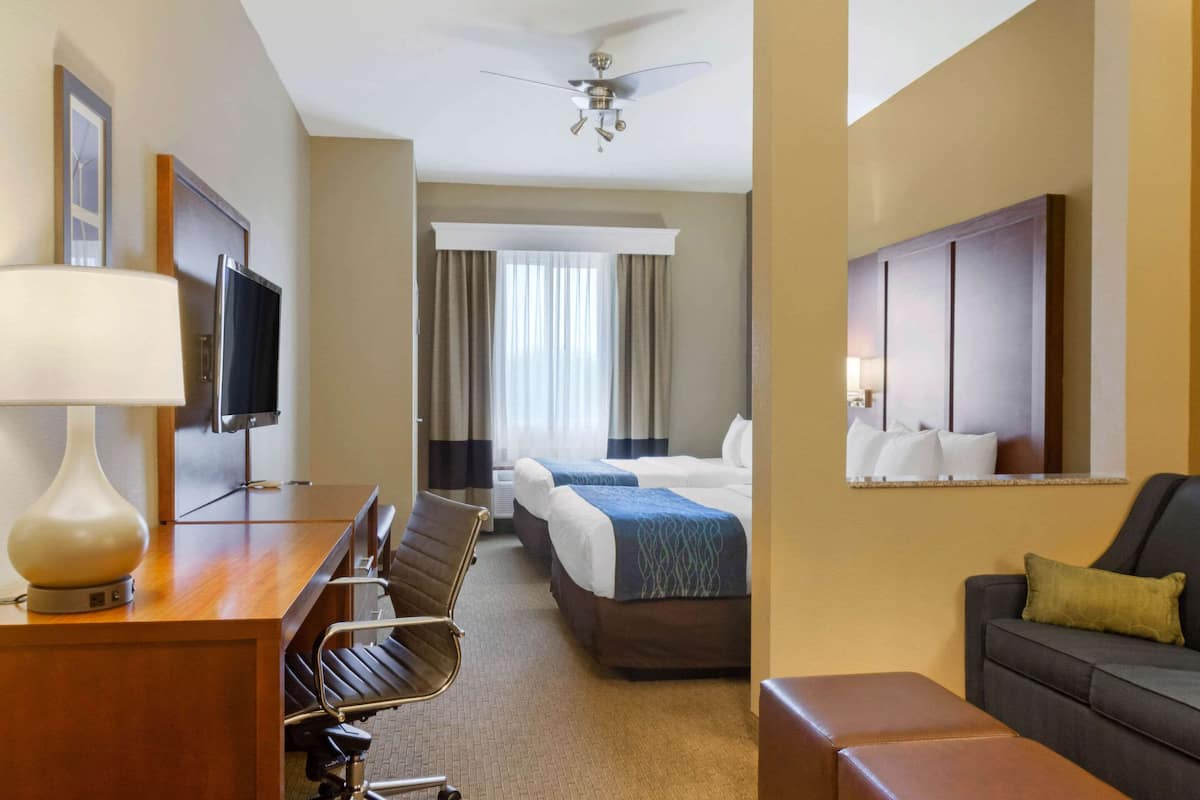 A hotel room with a desk, two beds, a sofa, a TV, and a ceiling fan.