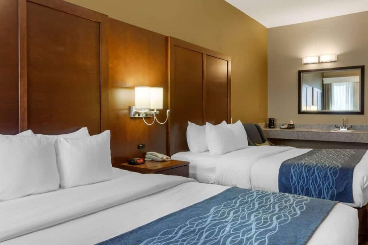 A hotel room with two beds, a nightstand with a phone, a lamp, a mirror, and a bathroom counter with a sink and mirror.