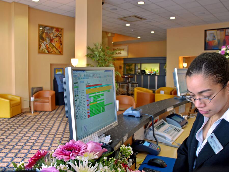 Una reception con una scrivania, un monitor computer, un telefono e un cartellino identificativo. Si notano sedie e una pianta sullo sfondo.