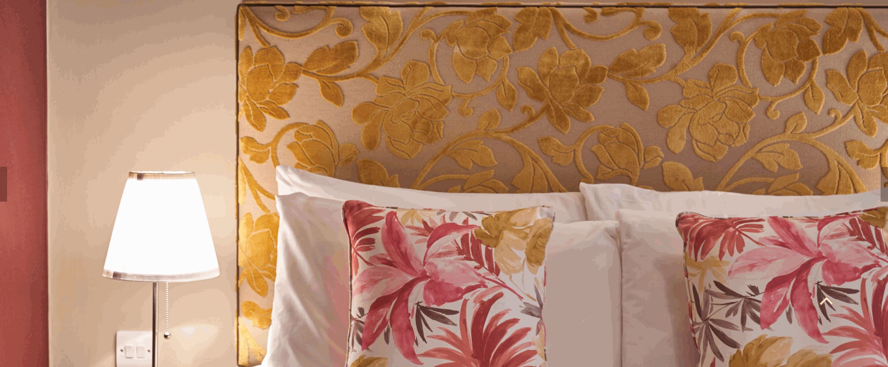A hotel room with a floral-patterned headboard, a bedside lamp, and floral-patterned pillows.