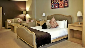 Quarto de hotel com uma cama grande, duas poltronas, um sofá, uma mesa de cabeceira de madeira e uma obra de arte na parede.