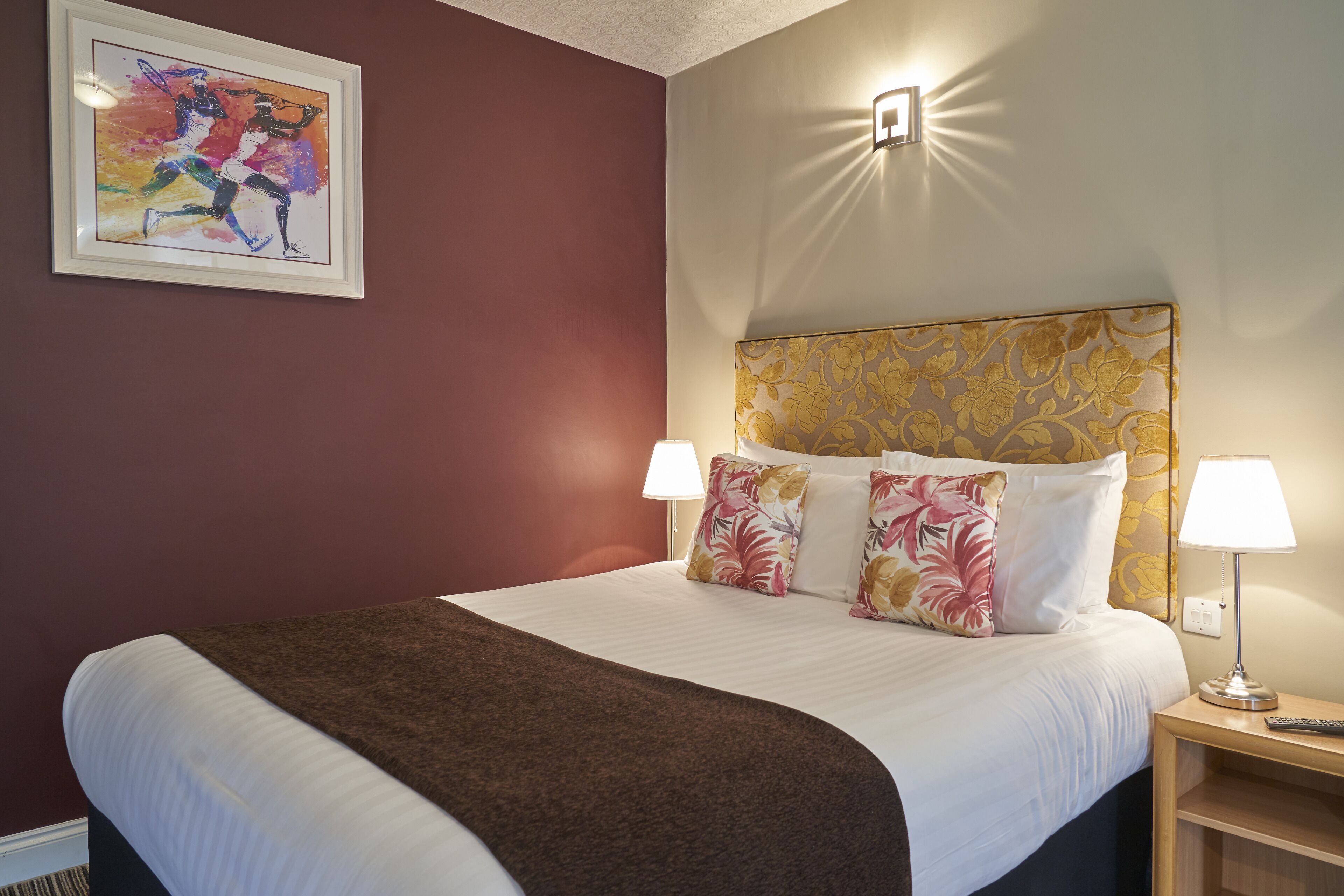 Une chambre d’hôtel avec un grand lit, deux lampes de chevet, une tête de lit ornée de motifs floraux et une œuvre d’art encadrée accrochée au mur.
