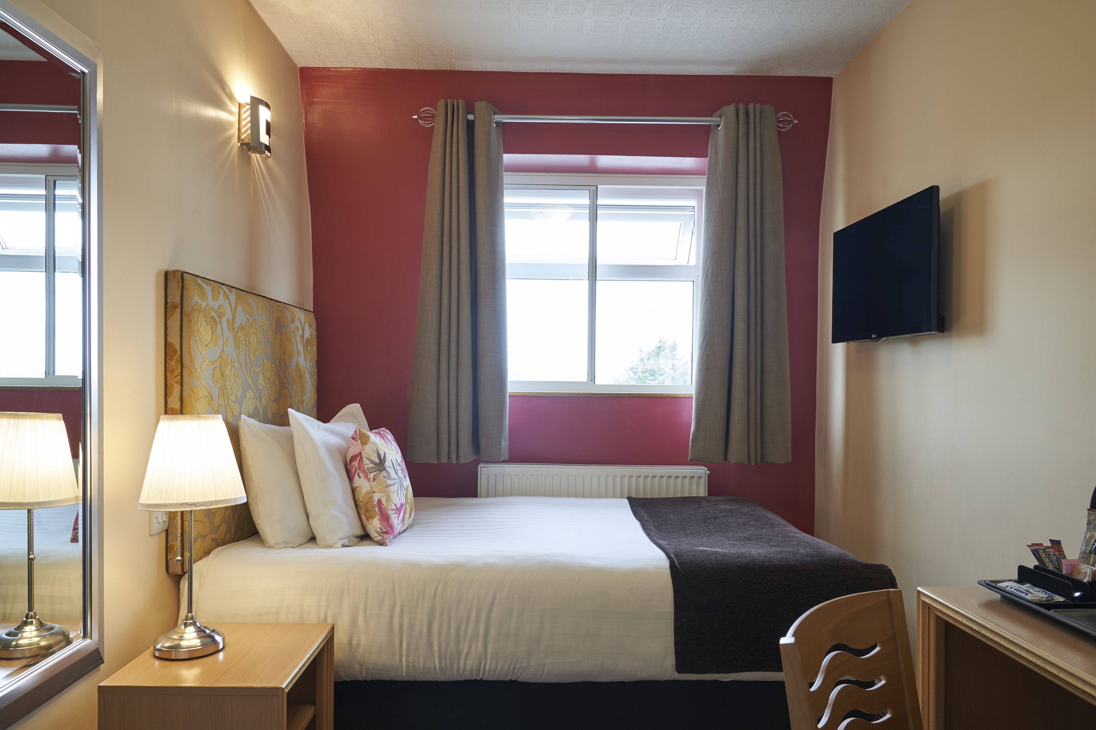 A hotel room with a bed, a desk, a lamp, a TV, and a window with curtains.