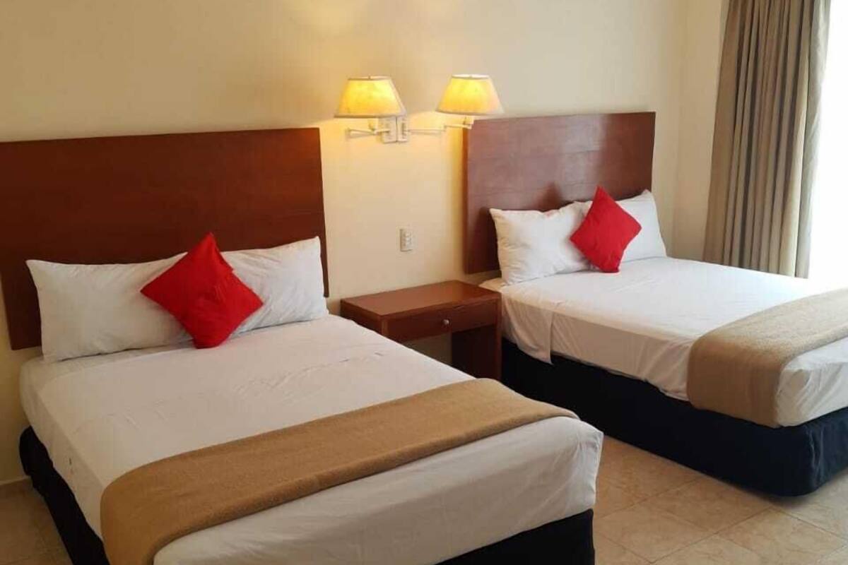 Deux lits jumeaux avec des têtes de lit, chacun agrémenté d’un oreiller rouge, d’une table de chevet en bois et d’une lampe fixée au mur.