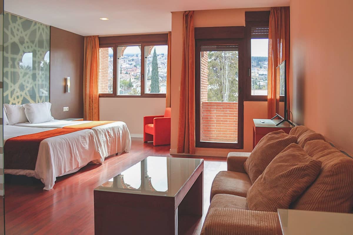 A hotel room with a bed, a sofa, a coffee table, a desk, and a view of the city.