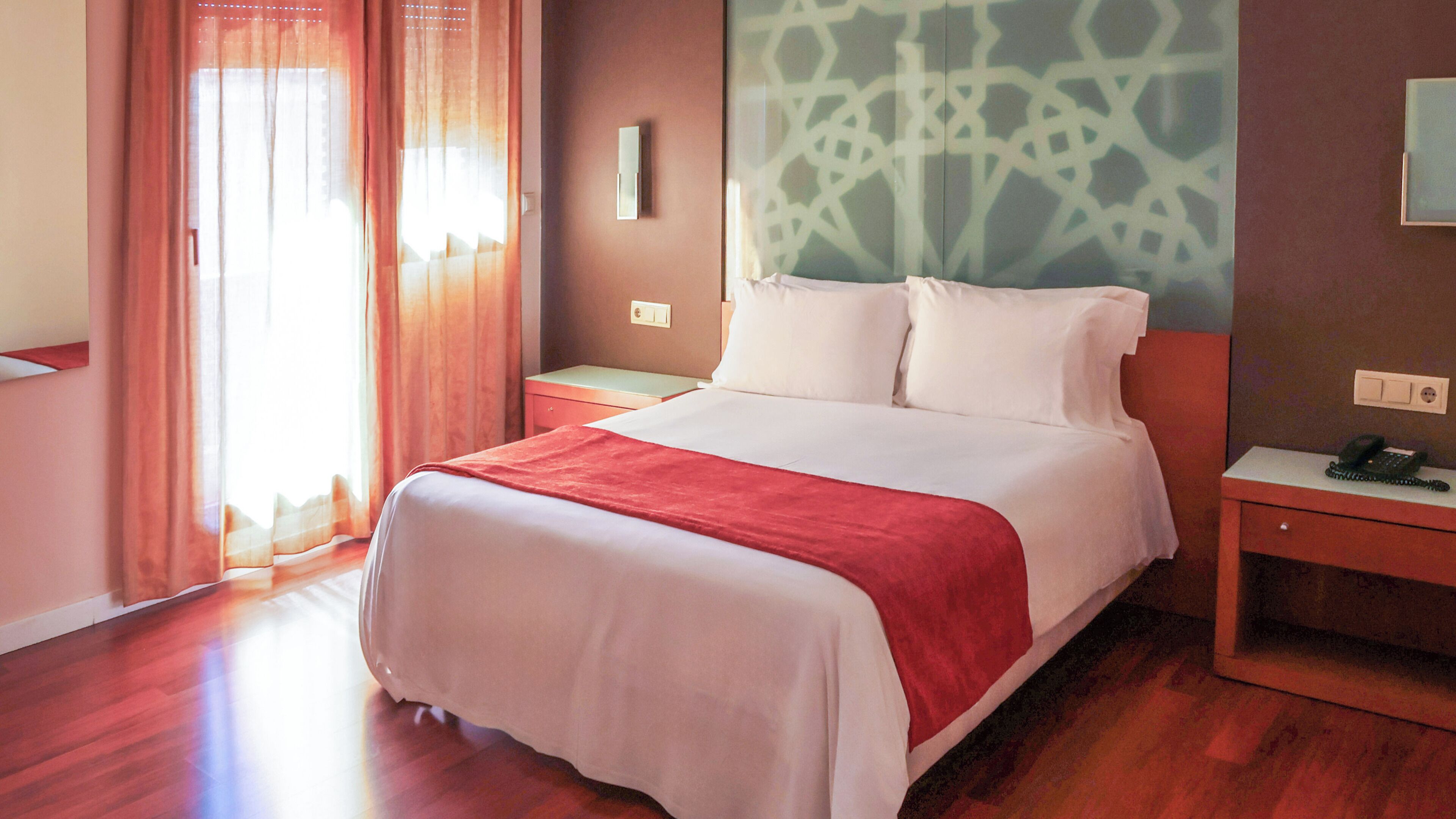 A hotel room with a bed, a nightstand with a phone, a wall-mounted light, and a window with curtains.