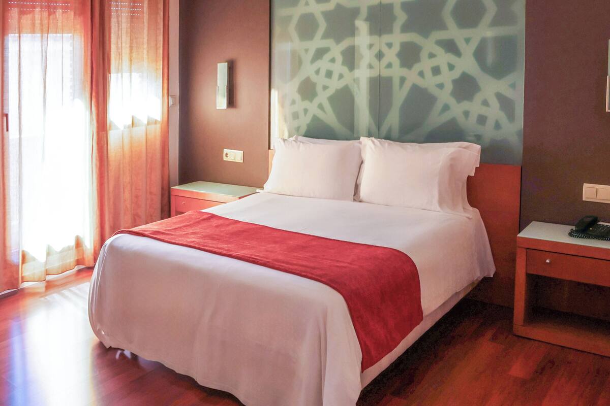 A hotel room with a bed, a nightstand with a phone, a wall-mounted light, and a window with curtains.