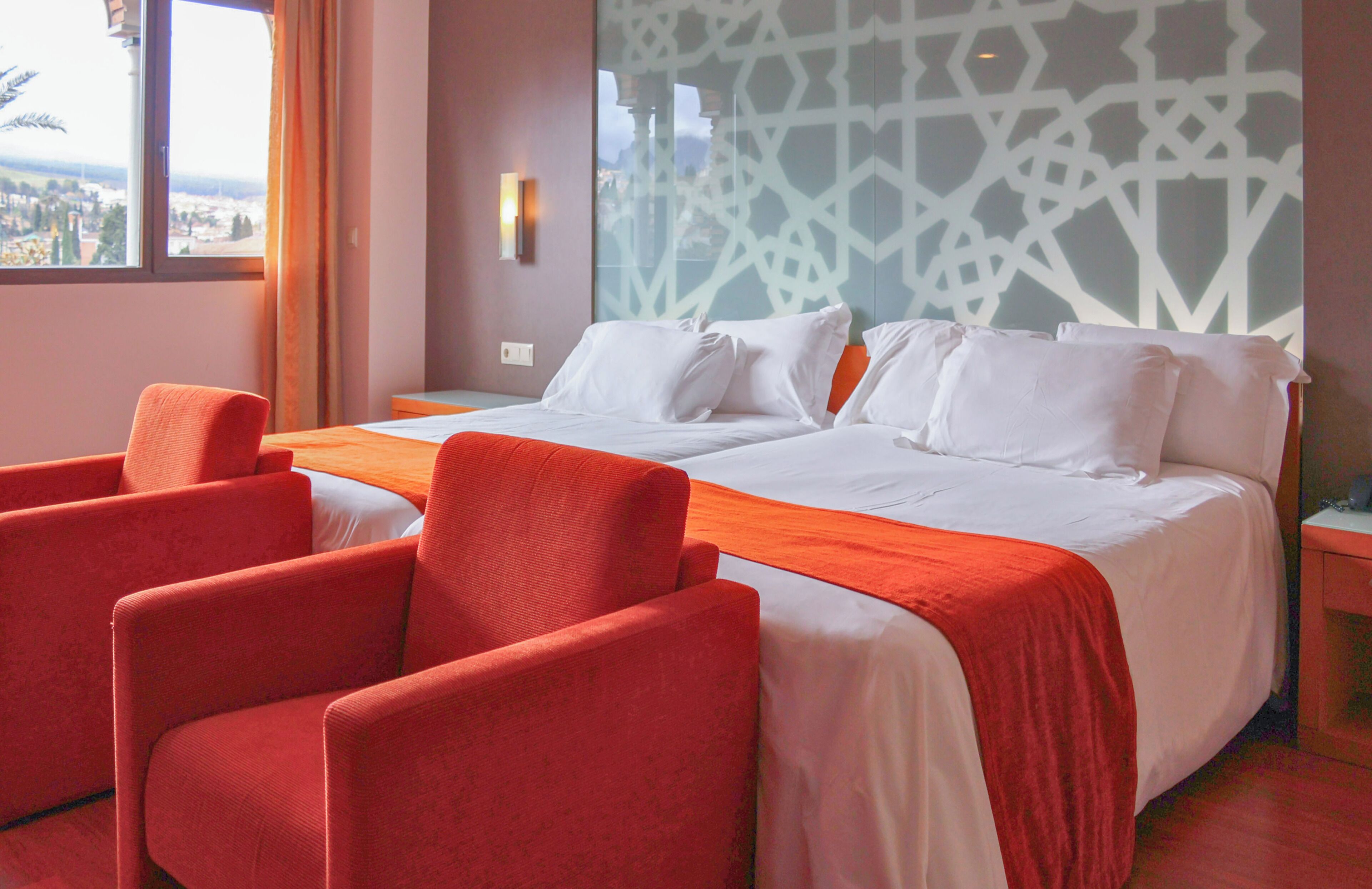 Habitación de hotel con una cama grande, dos sillones rojos, vistas a la ciudad y un papel tapiz estampado.