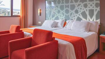 Habitación de hotel con una cama grande, dos sillones rojos, vistas a la ciudad y un papel tapiz estampado.