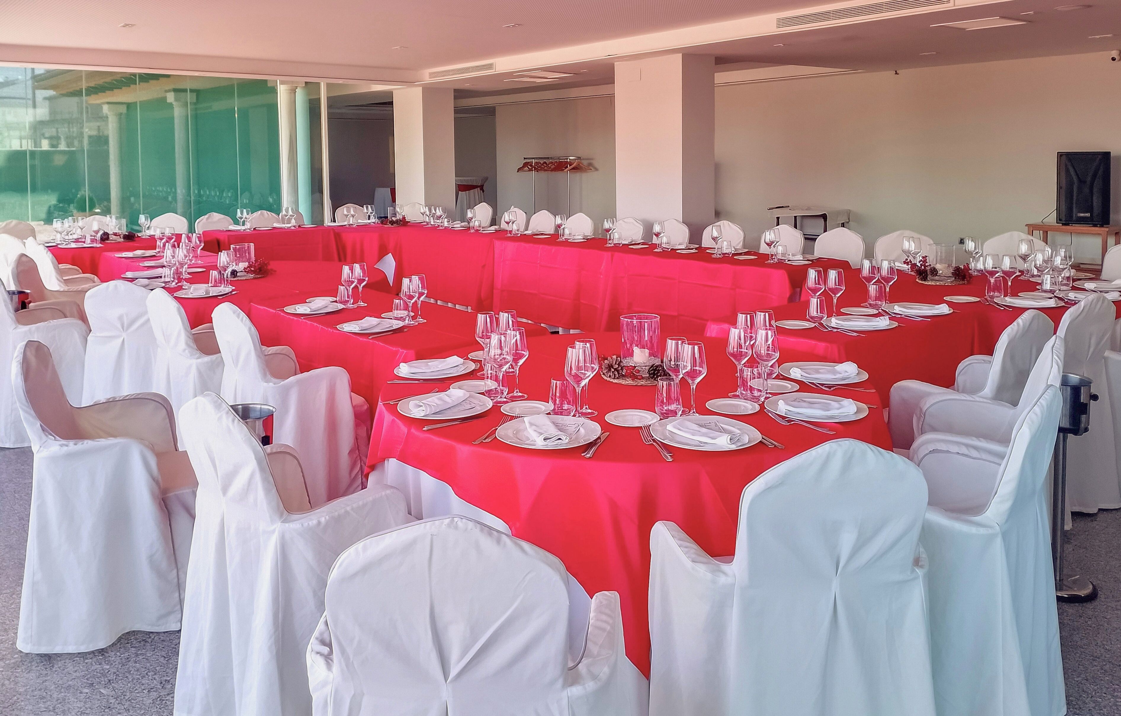 Un salón de banquetes con largas mesas dispuestas para un evento formal, sillas blancas y mantel rojo.