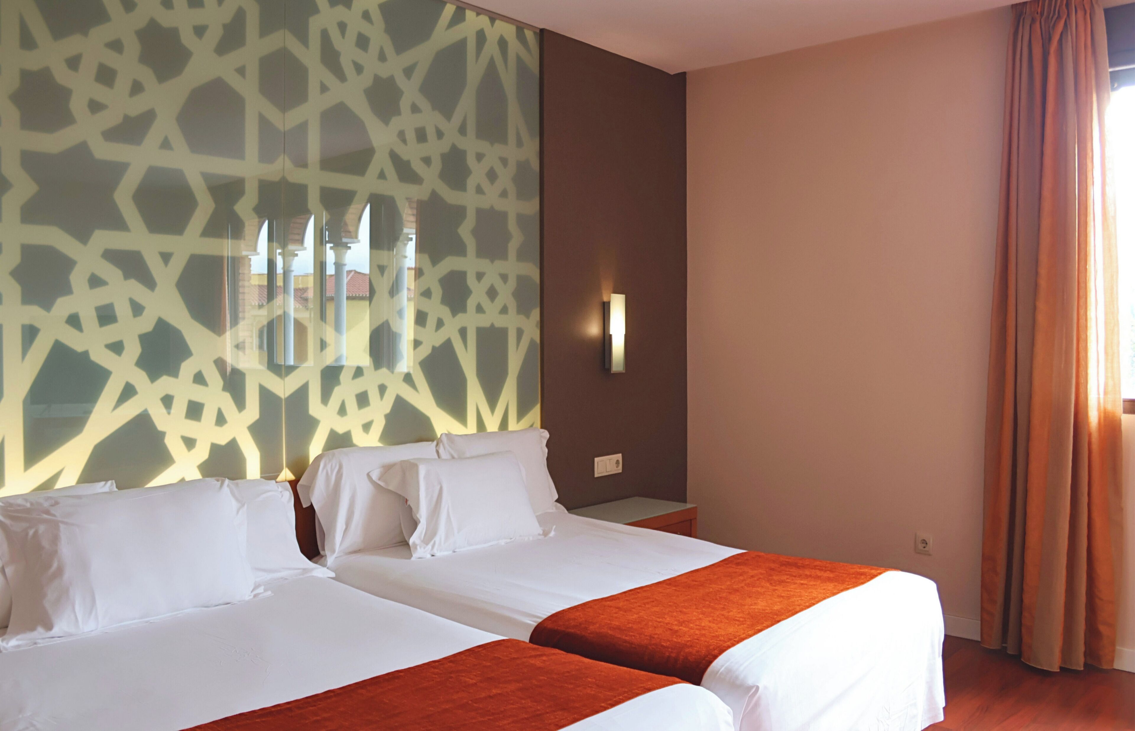 A hotel room with two beds, a patterned wall, a nightstand, and a window with curtains.