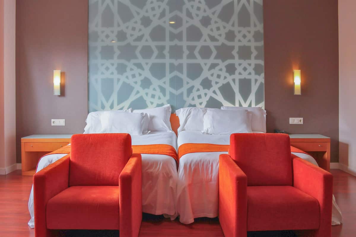 A hotel room with a large bed, two red armchairs, a nightstand, and a wall with a geometric pattern.
