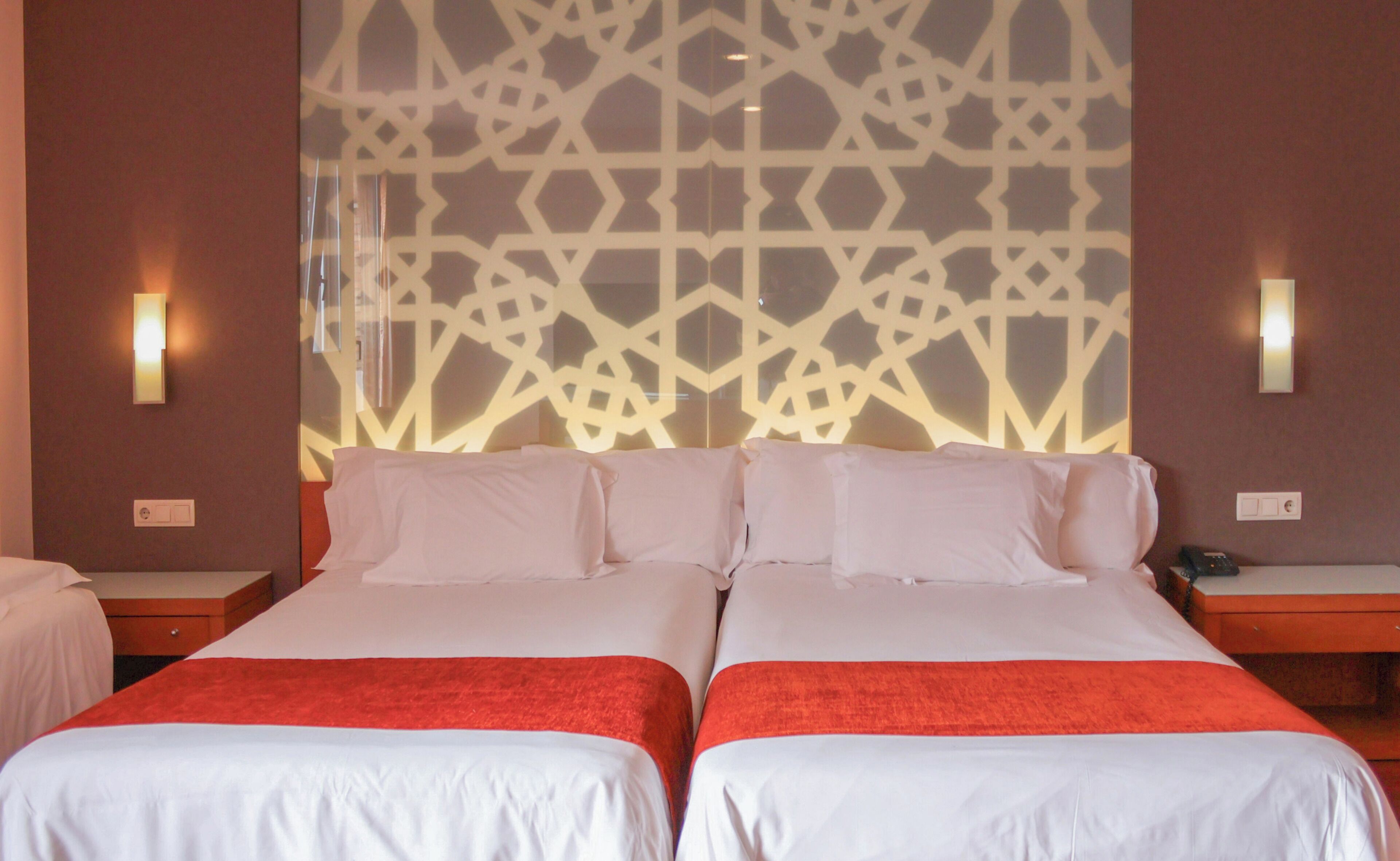 A hotel room with two beds, a patterned wall, and wall-mounted lights.