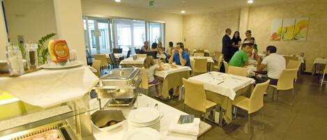 Un comedor tipo bufé con mesas y sillas, un mostrador de bufé con comida, y un grupo de personas cenando.