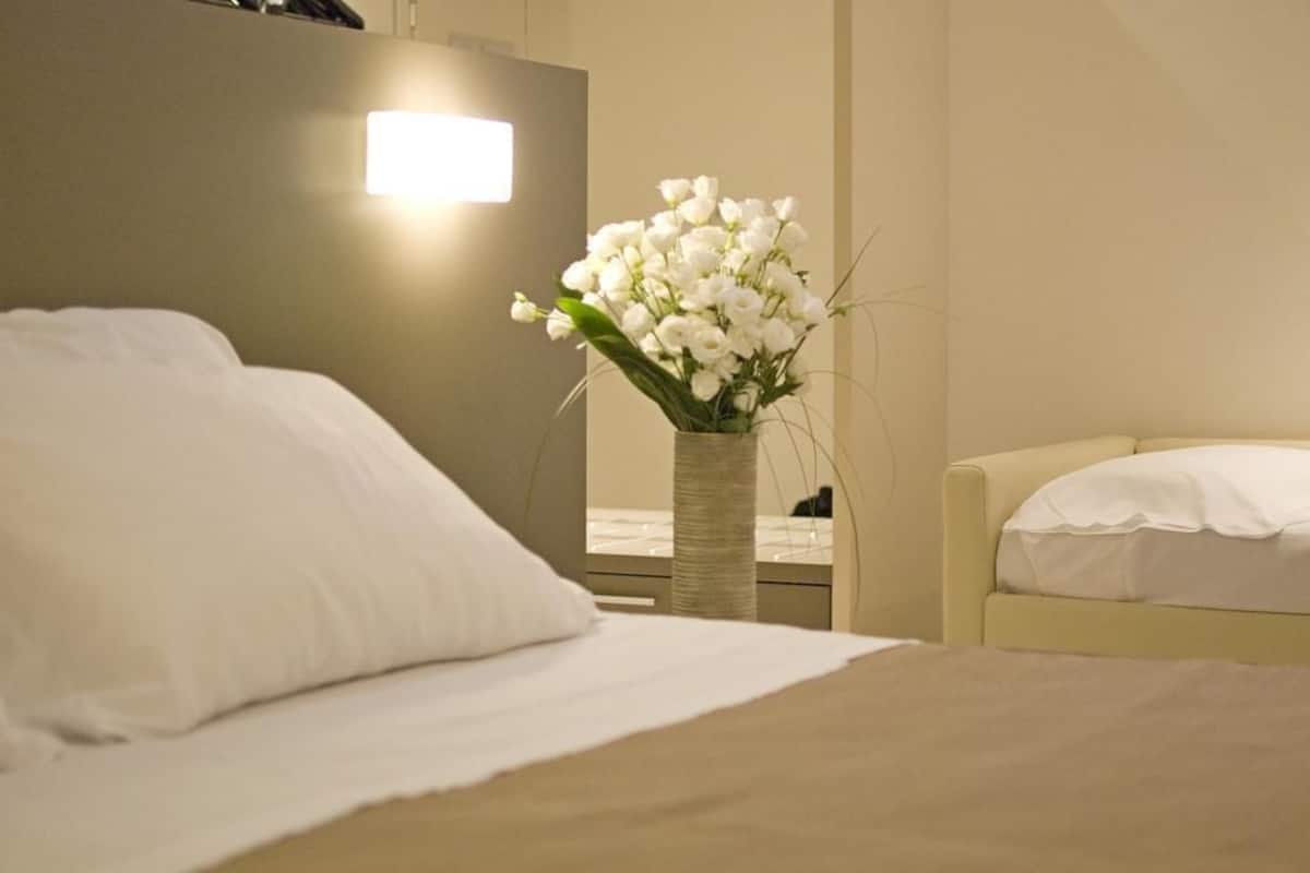 A hotel room with a bed, a bedside table, a vase with white flowers, and a lamp.