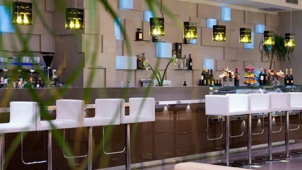 A modern bar area with white bar stools, a counter with bottles, and hanging light fixtures. - Hotel Cube (Ravenna)