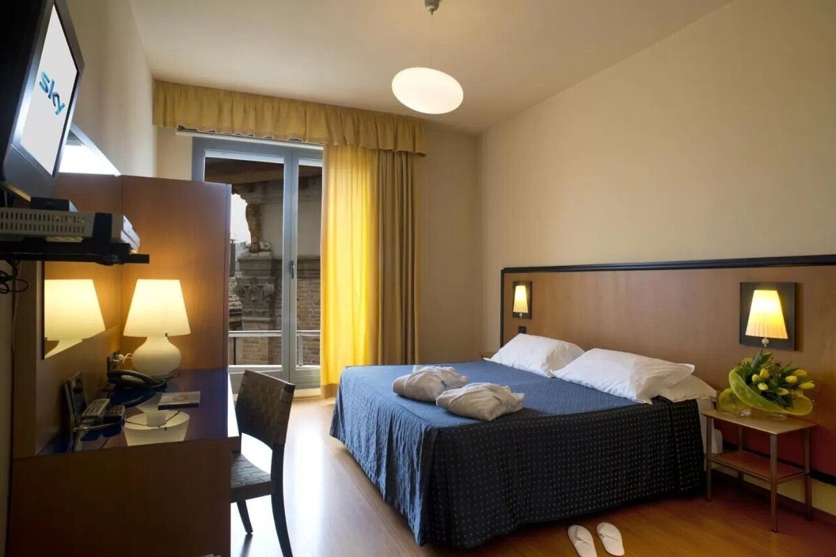 A hotel room with a bed, a desk with a telephone, a chair, a small table with a lamp, and a wall-mounted TV.