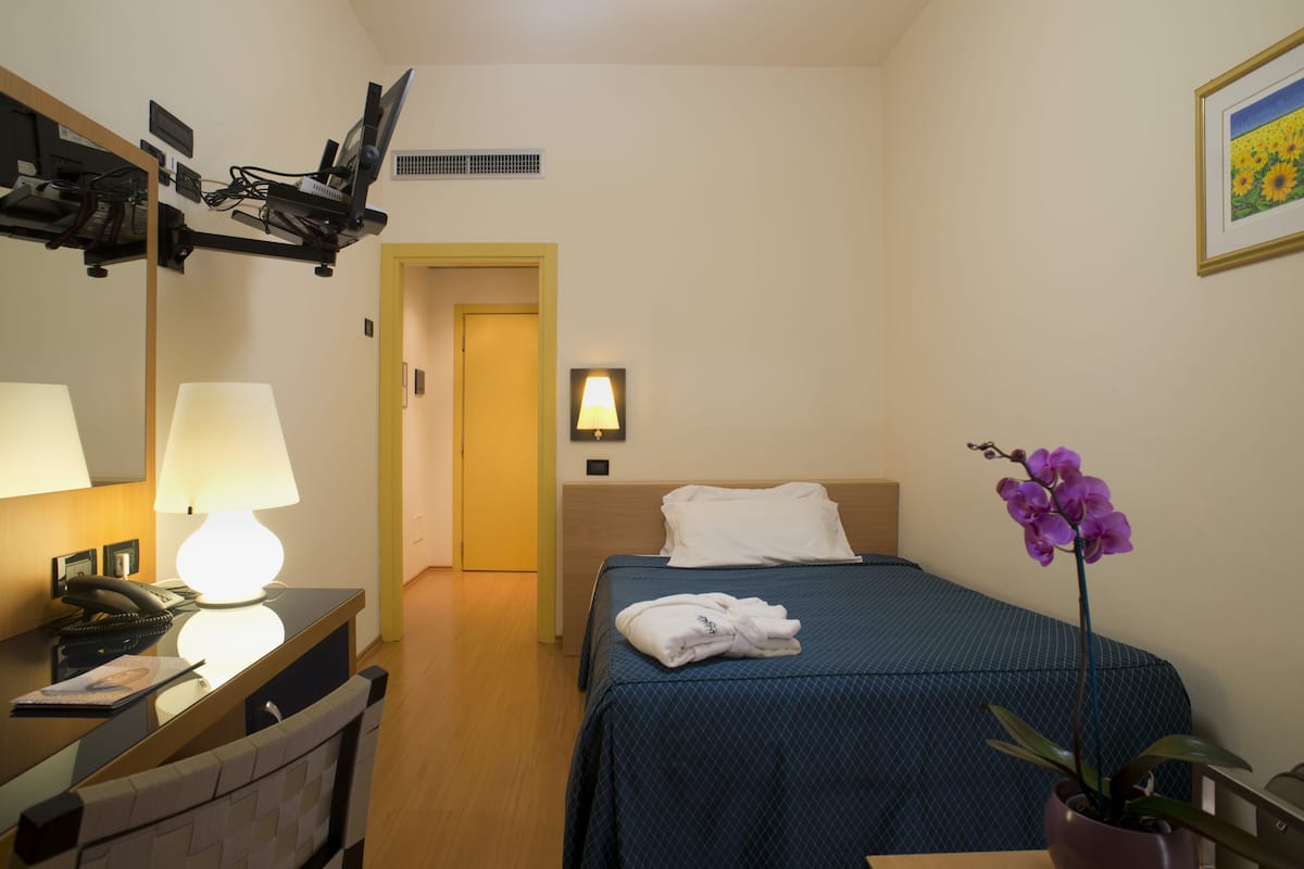 A hotel room with a bed, a desk with a lamp, a telephone, a mirror, a wall-mounted TV, and a potted orchid.