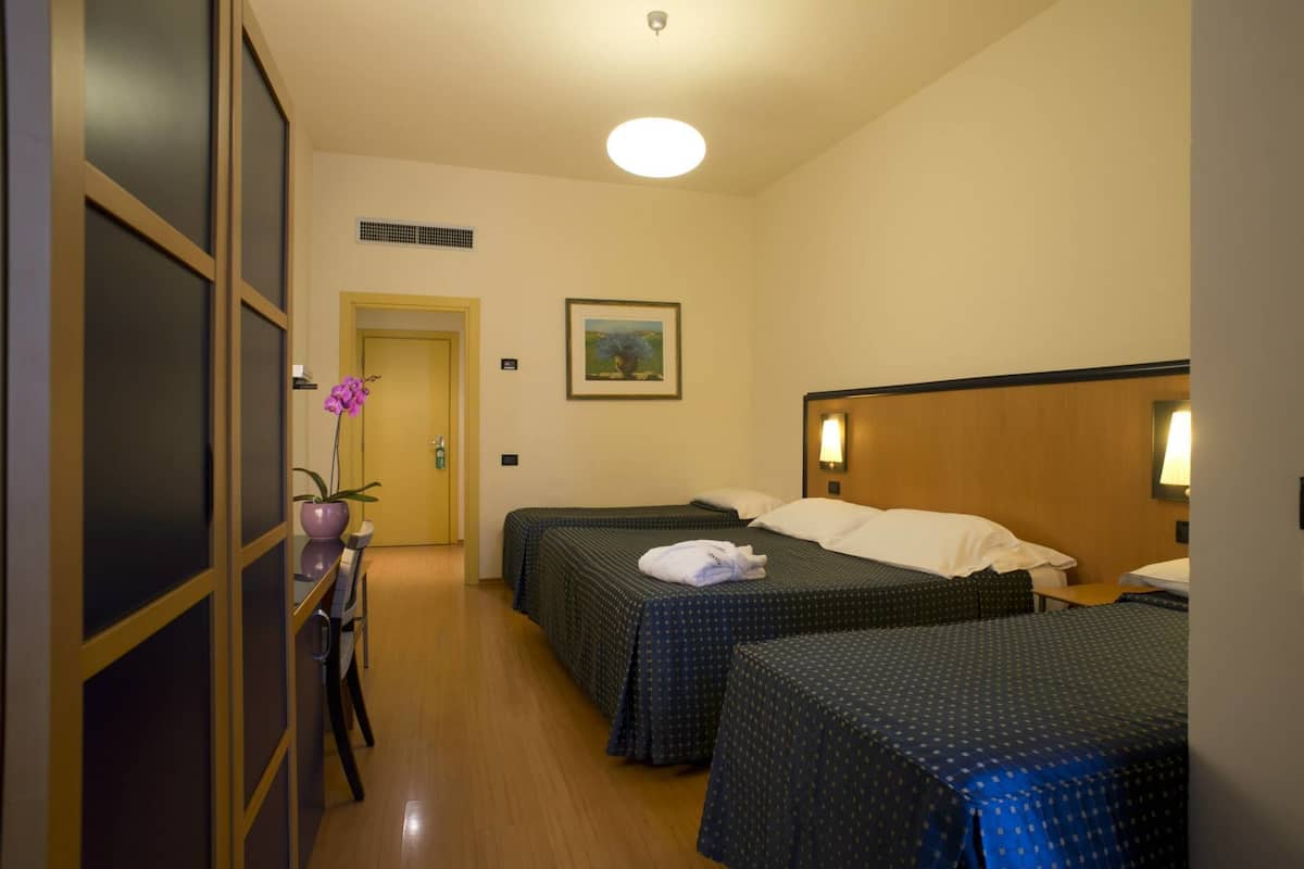 A hotel room with two beds, a desk, a chair, a potted plant, and a painting on the wall.