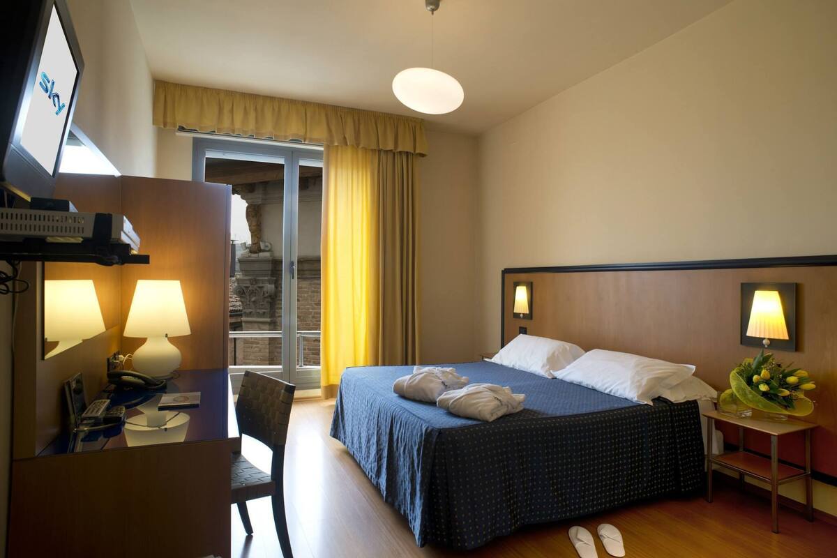 A hotel room with a bed, a desk with a telephone, a chair, a lamp, and a small table with a flower arrangement.