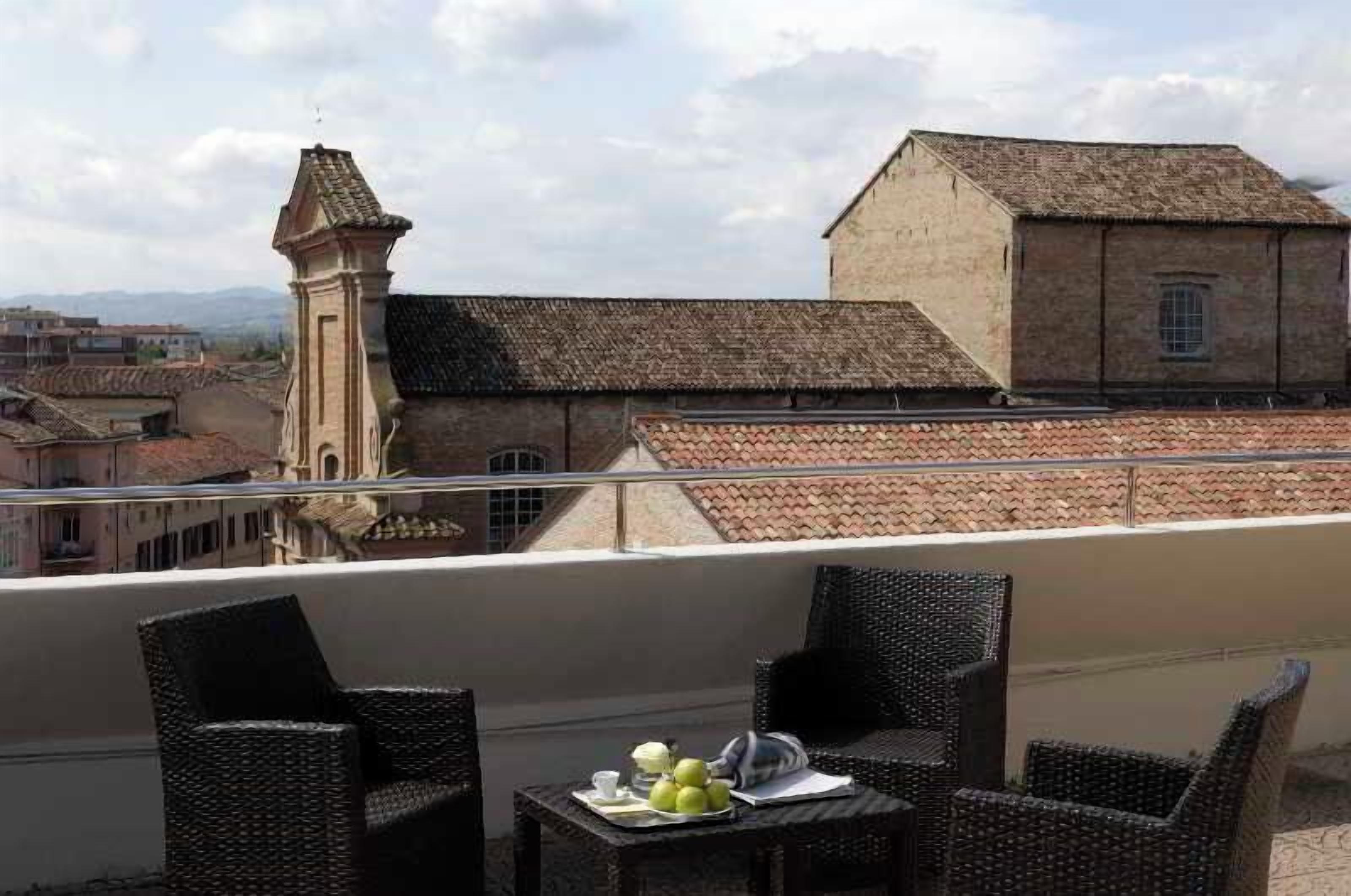 Un balcone con mobili in vimini e un tavolo con frutta, vista su un edificio storico con una torre.