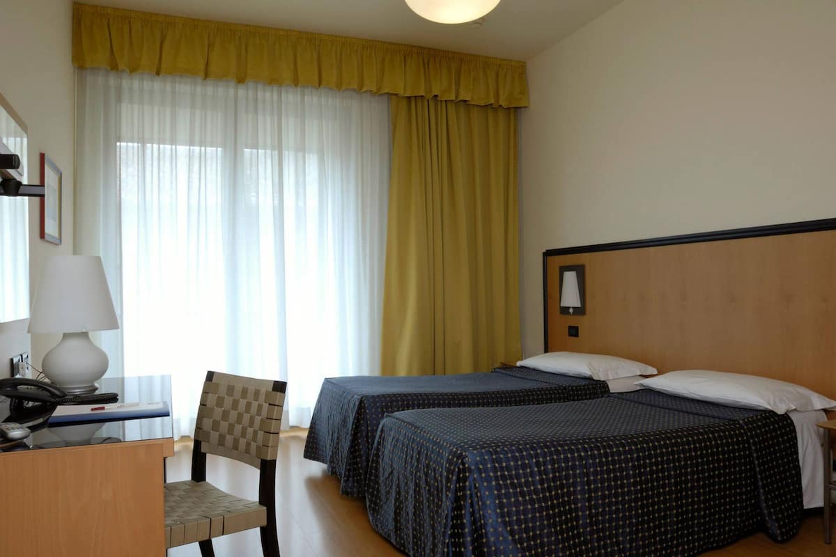 A hotel room with two beds, a desk with a lamp, a chair, and a large window with curtains.