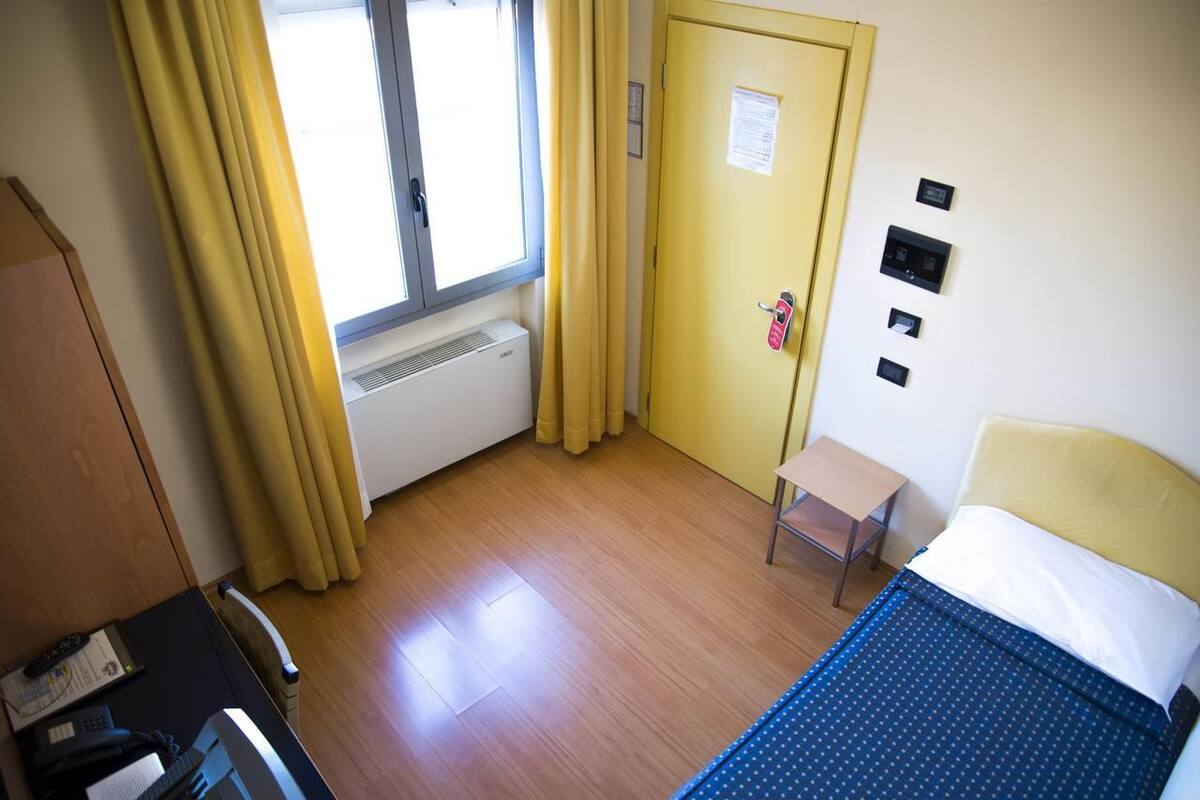 A hotel room with a bed, a desk, a chair, a window with curtains, a radiator, and a door with a lock and a keycard reader.
