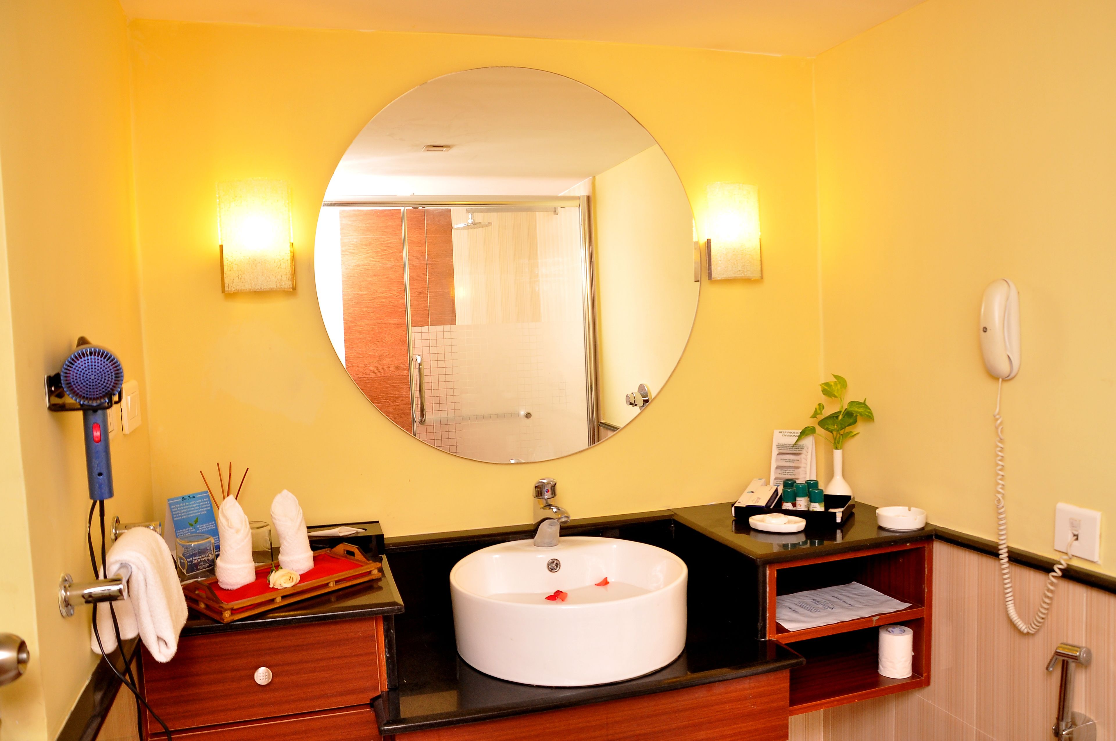A bathroom with a round mirror, a white sink, a towel rack, a hairdryer, and a telephone.