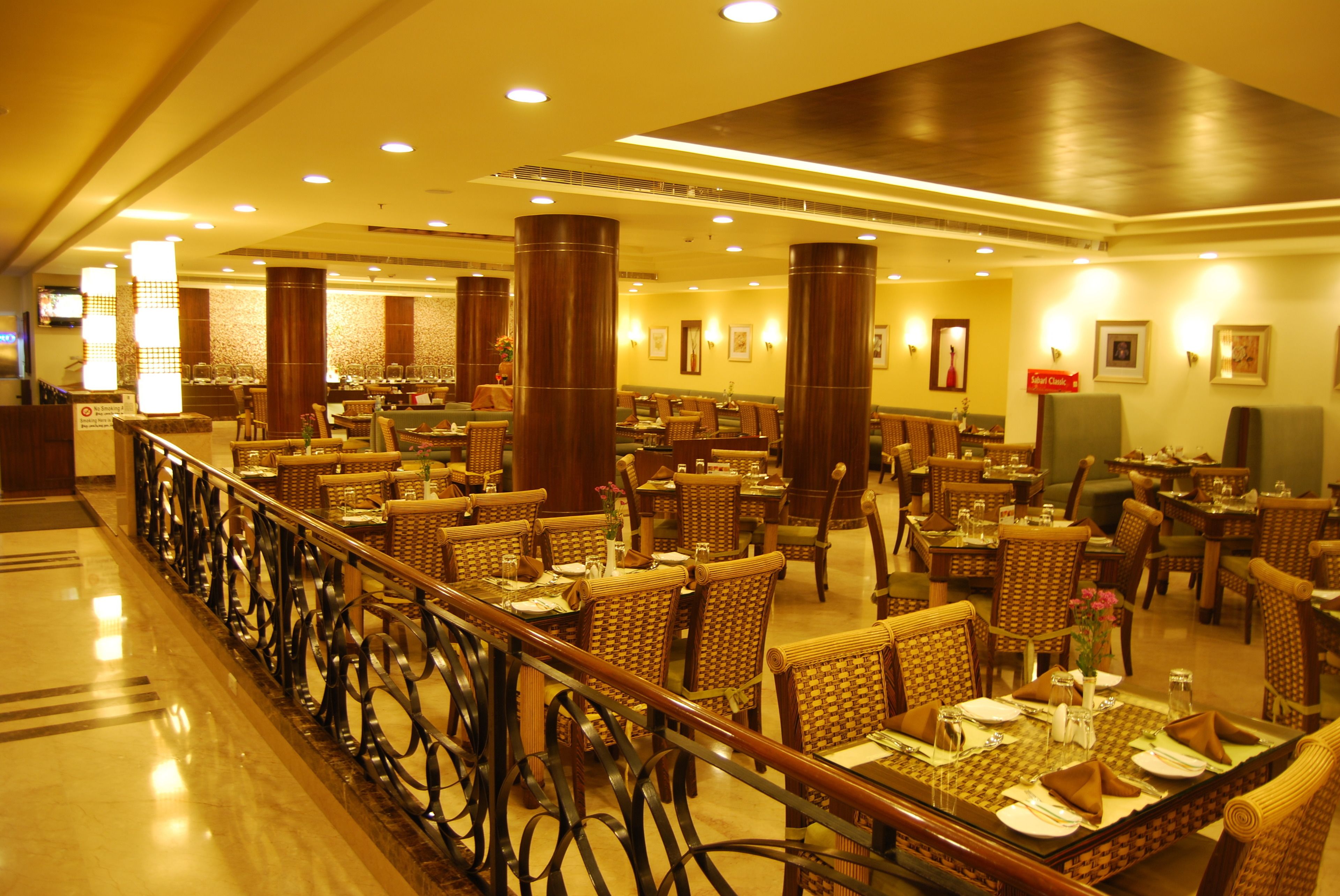 A spacious hotel dining area with multiple tables and wicker chairs, featuring a polished floor and a railing along the walkway.