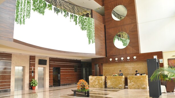 A modern hotel lobby with a high ceiling, wooden paneling, marble flooring, reception desks, elevators, and a large window allowing natural light. - Gokulam Park Sabari OMR Hotel (Tirupporur)