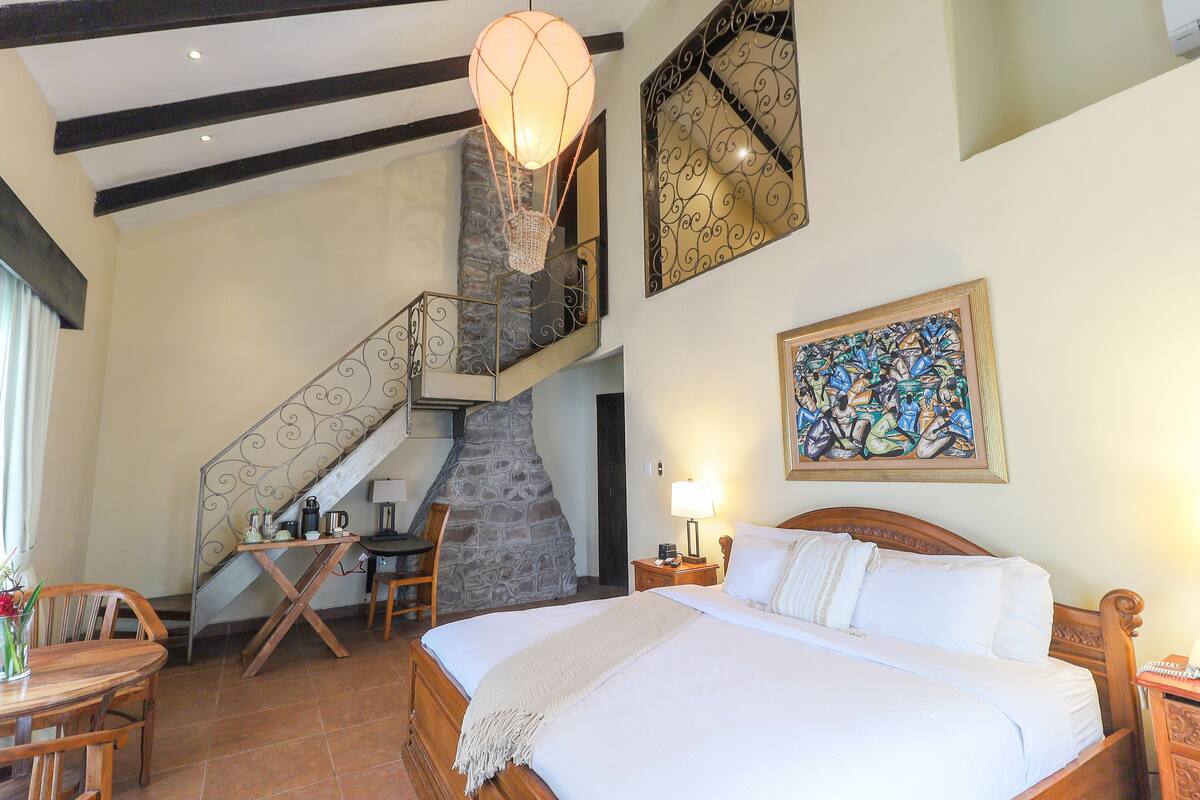 A bedroom with a large bed, a stone fireplace, a staircase, and a dining area.