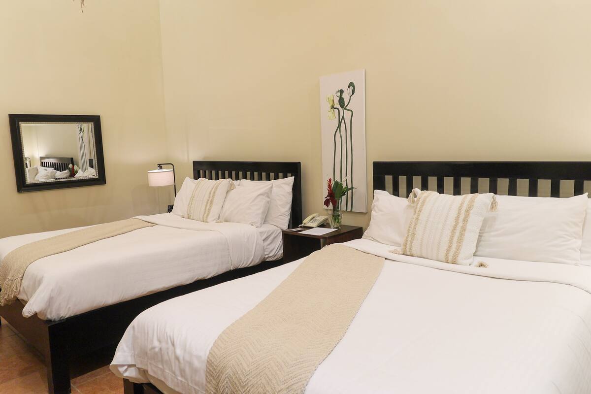 A hotel room with two beds, a bedside table with a lamp, a mirror, and a wall-mounted artwork.