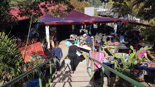 A market with various stalls selling goods, a staircase leading to an upper level, and a red-roofed building in the background. - The Riverside Inn (Los Naranjos)