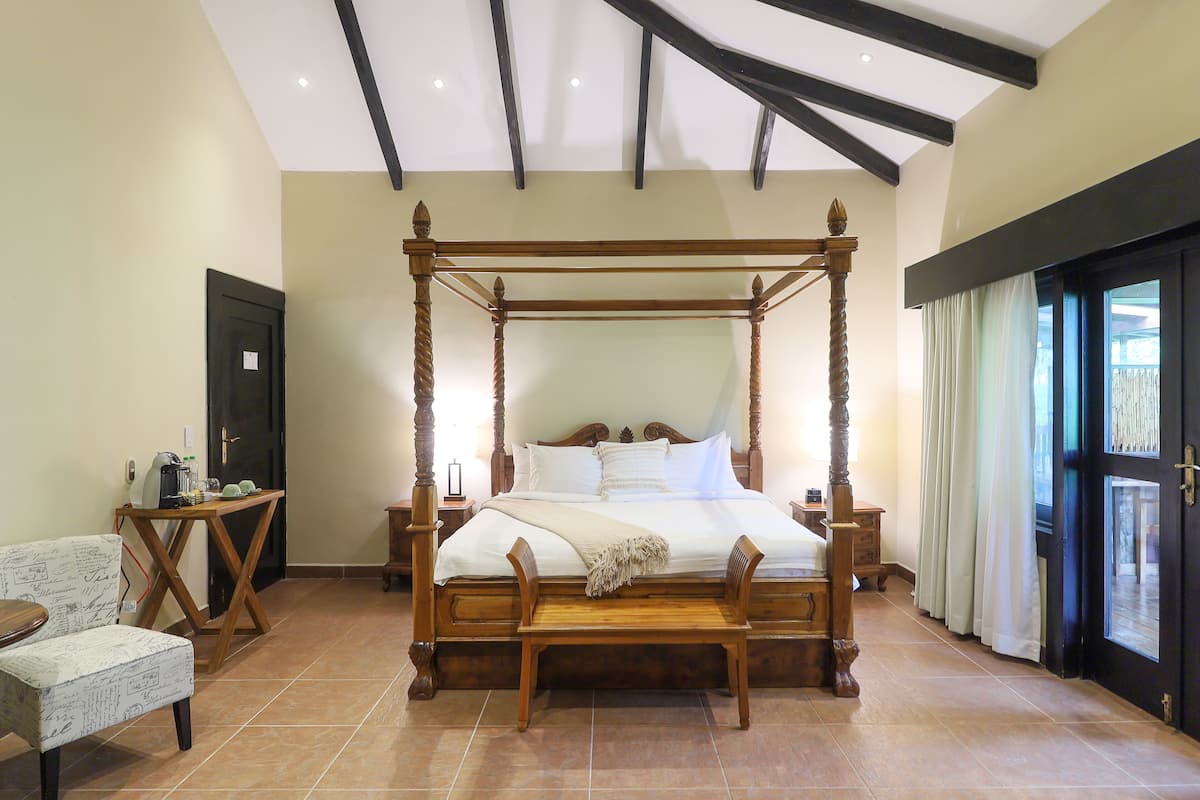 A four-poster bed with a wooden frame, a bedside table, a chair, and a window with curtains.