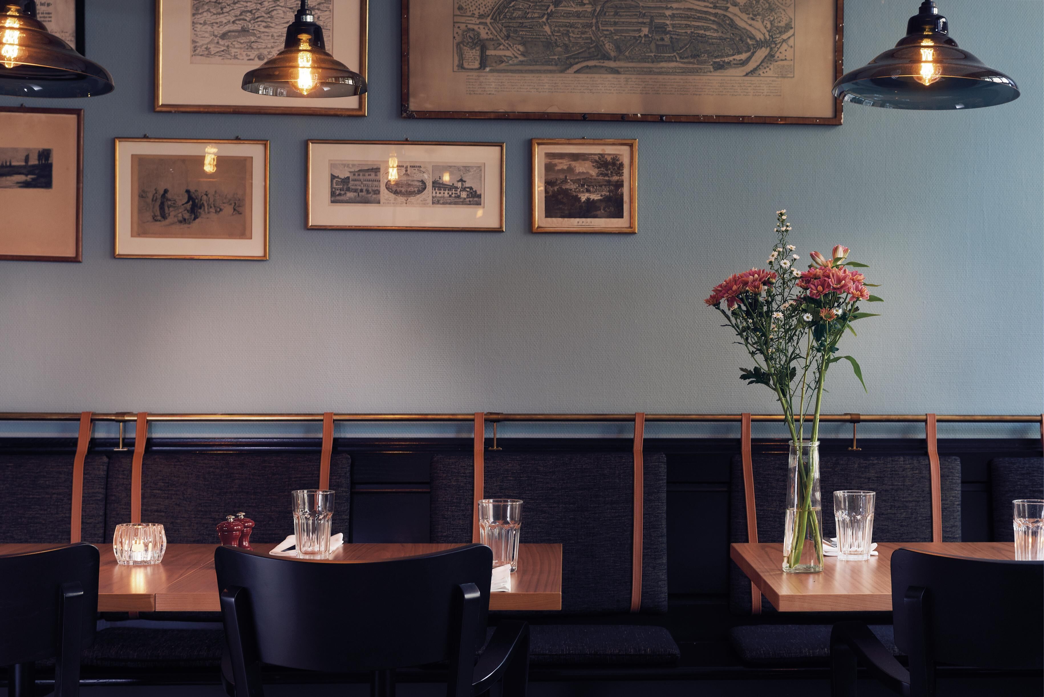 L’intérieur d’un restaurant, avec des tables, des chaises, des tableaux encadrés accrochés au mur et un vase rempli de fleurs posé sur une table.