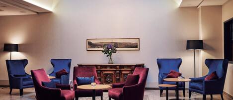 Un hall d’entrée avec quatre fauteuils bleus, deux tables rondes, un buffet en bois et un tableau représentant un paysage, encadré, accroché au mur.