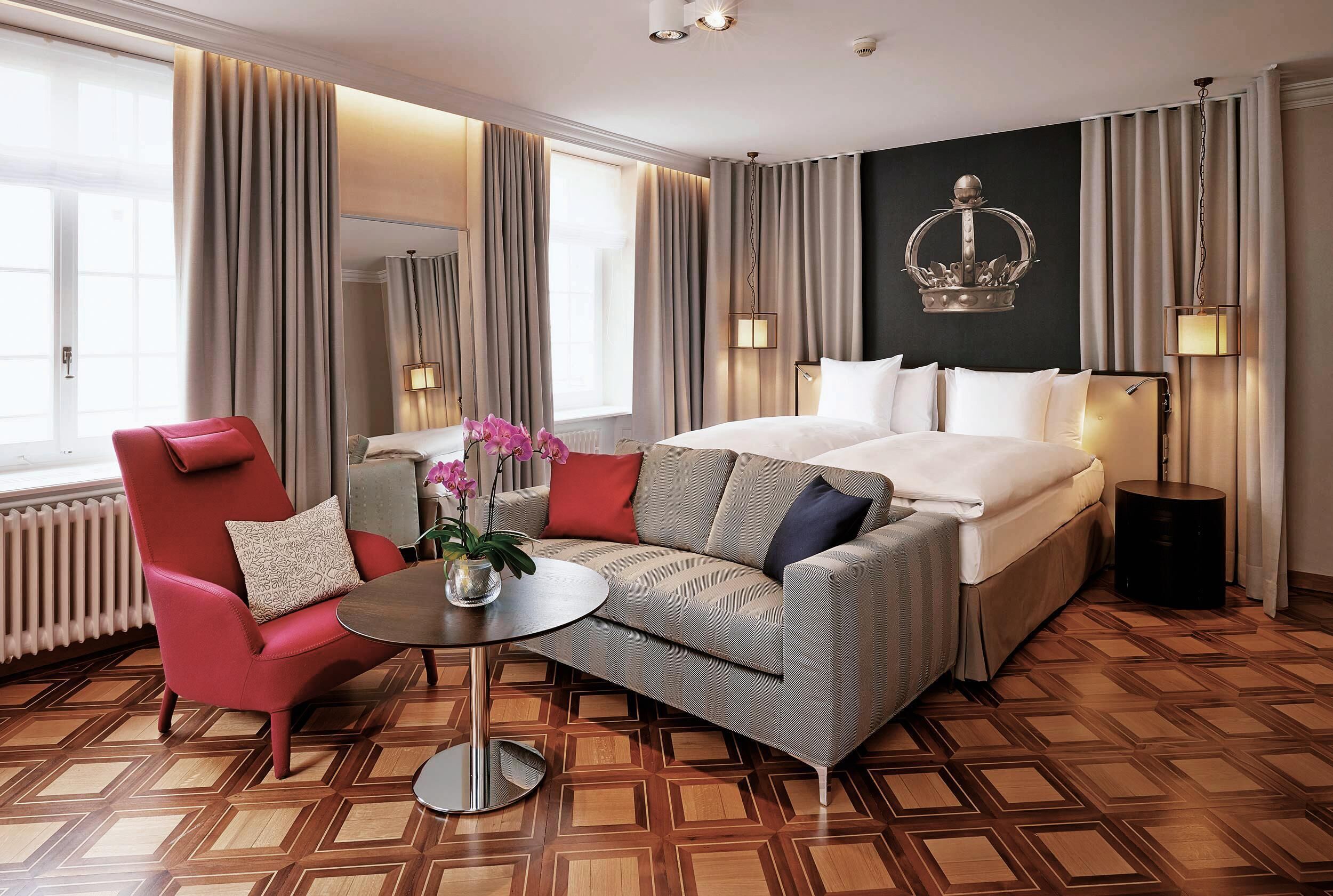 A hotel room with a bed, a red armchair, a grey sofa, a round table, and a decorative crown on the wall.